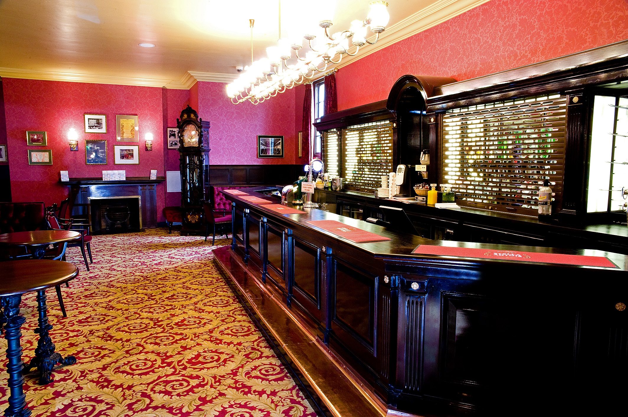Inside of the City Varieties bar. There is a dark wood bar on the right hand side, an antique grandfather clock in the distance and dark wood round tables on the left. The carpet is red with a gold pattern and the walls are also red.