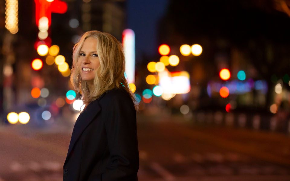 Vonda Shepard smiling, she is outside at night with glowing street lights behind her