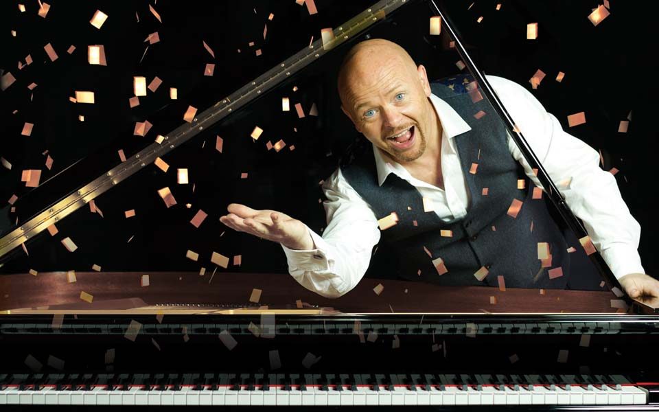 Comedian Jon Courtenay smiling under the lid of a grand piano with confetti falling