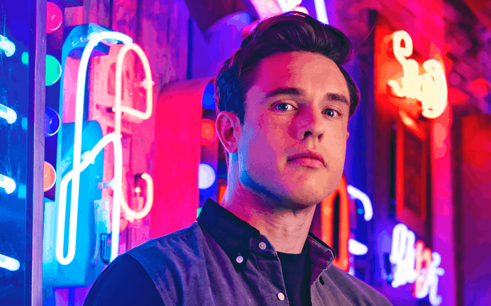Ed Gamble Ed Gamble looks into the camera whilst standing in front of some neon signs.
