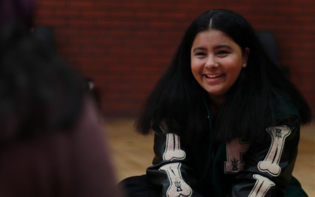HannahKerryMaule Young girl with long dark hair sat on the floor smiling.