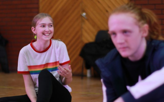 A girl with a rainbow top on smiling with another young person blurry in the foreground.
