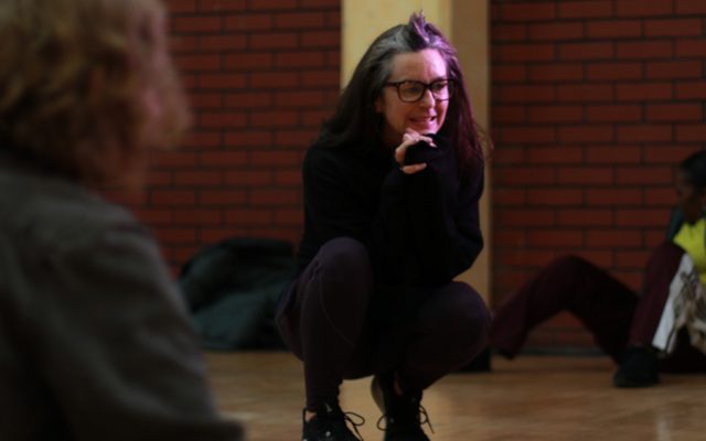 Lizi Patch. Credit Kerry Maule A woman crouching on the floor, dressed in black with her hand to her face.