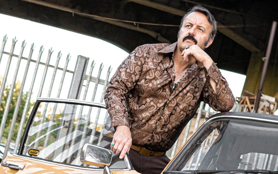 Comedian Mike Bubbins wearing a patterned shirt and leaning on a vintage car.