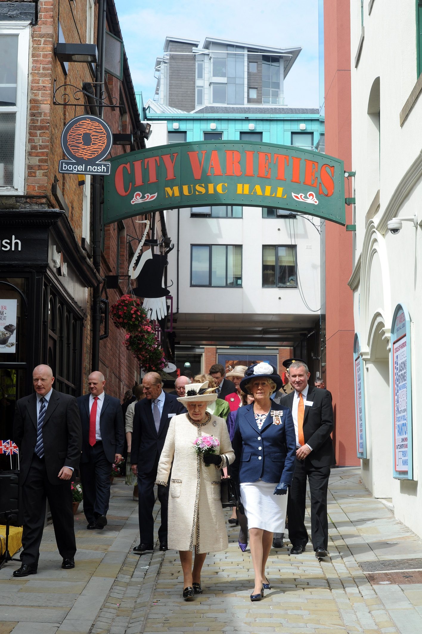 The Queen wearing a cream dress and hat followed by a group of people in suits walked down a narrow road. The sign above her reads City Varieties Music Hall.