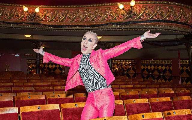 ETAJ. Jamie Campbell auditorium-min Jamie Campbell in a pink jacket and trousers stands with his arms outstretched in the theatre auditorium