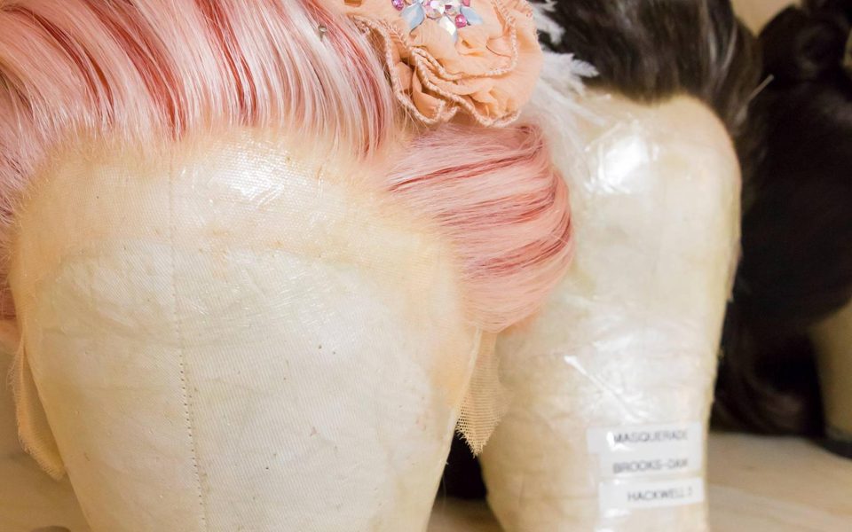 A pale pink streaky wig on a wig stand with a flower pinned into it
