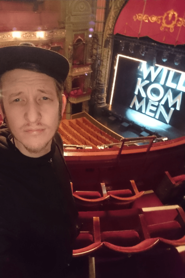 Adam Norton stands in Leeds Grand Theatre's balcony with the stage behind him showing the Cabaret sign Wilkommen in large white letters