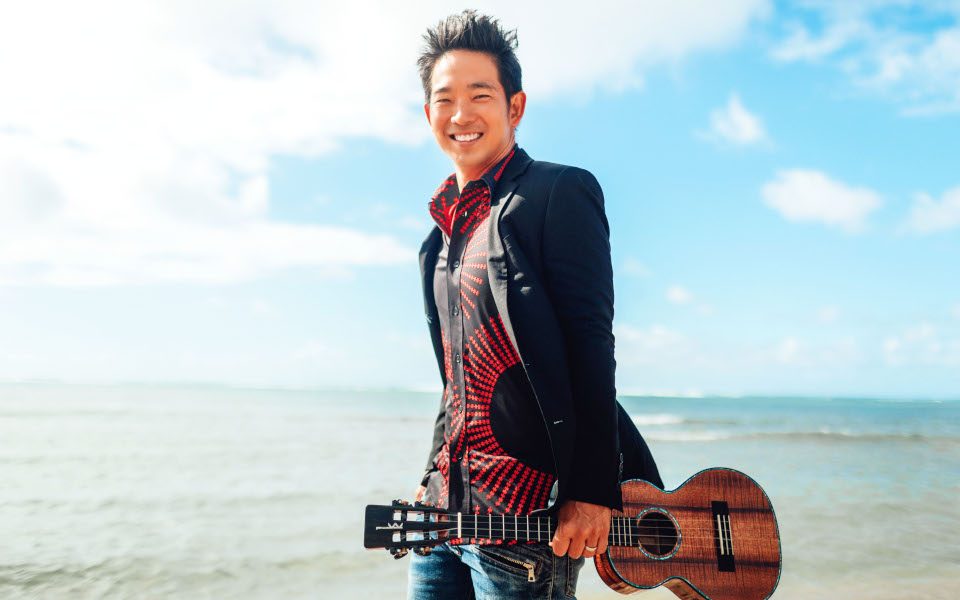 Jake Shimabukuro on the beach with his ukulele smiling