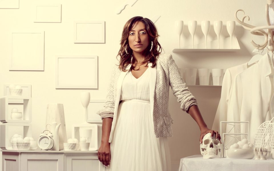 Shazia Mirza in a white dress against a white background and surrounded by various white things