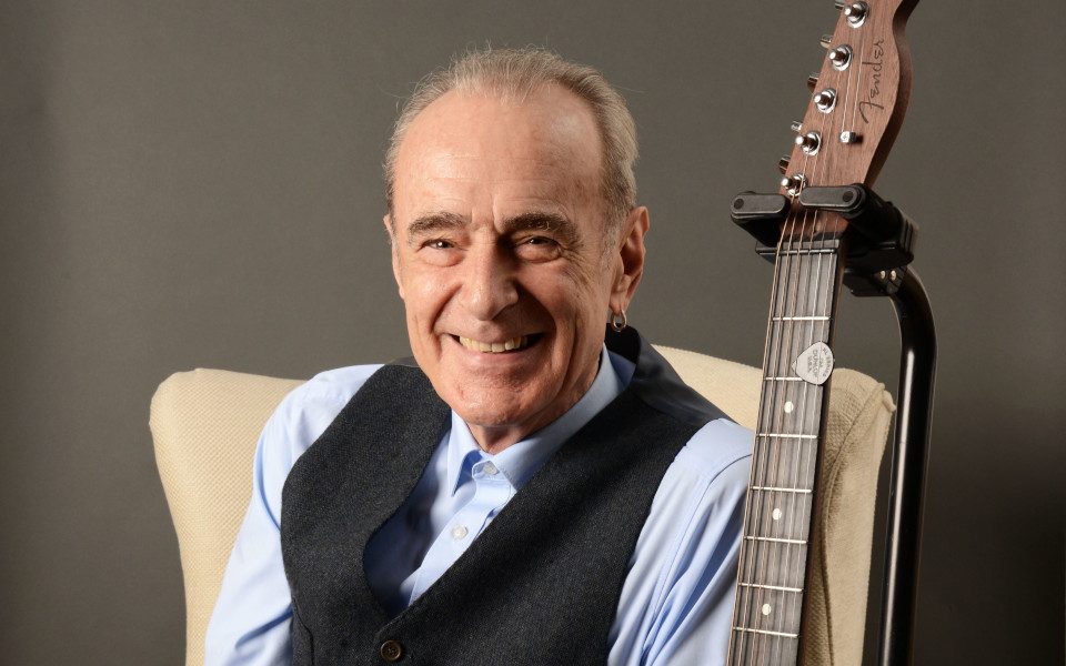 Francis Rossi smiling, wearing a waistcoat with an acoustic guitar