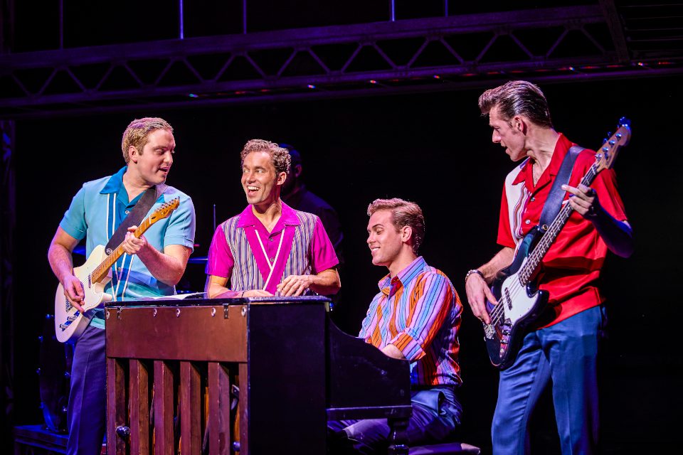 Four men around a piano. One plays it and two play guitar