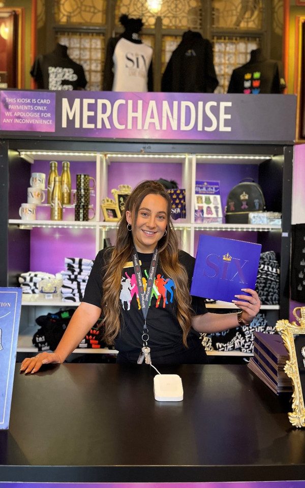 Jessica Davison holding up a SIX programme in front of the merchandise stand