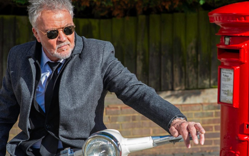 Paul Young sat on a Vespa scooter wearing shades and a suit by a red post box
