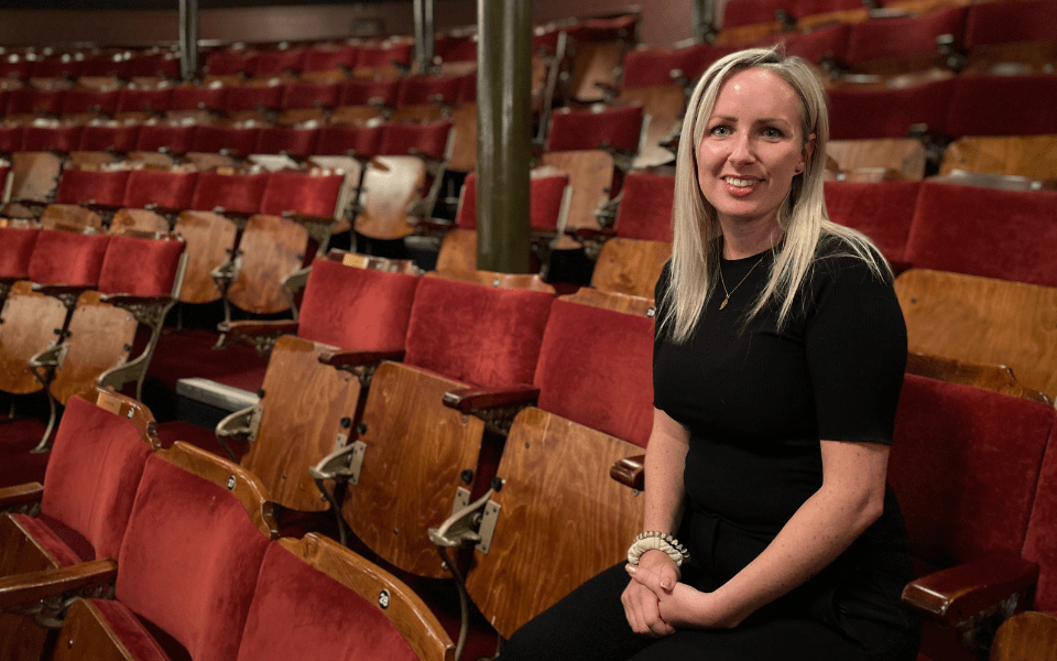 Kirsty Triplett, wearing a black t-shirt and trousers, smiles to camera whilst sat in the auditorium at Leeds Grand Theatre