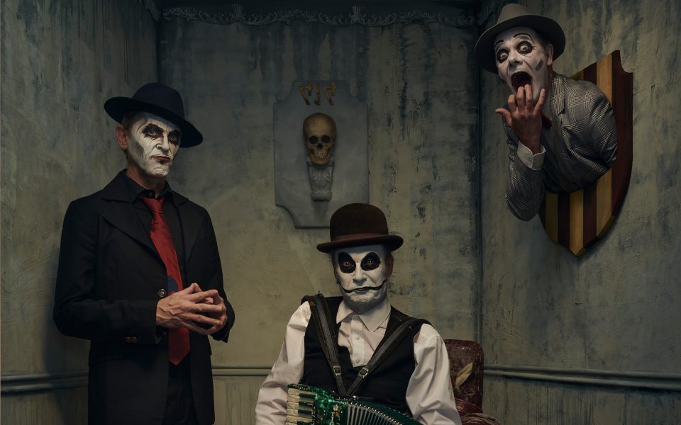The Tiger Lillies wearing their iconic white make-up and sat in a creepy dark room, one member has their head hanging on the wall like a hunters trophy, another is sat on a chair playing an accordion
