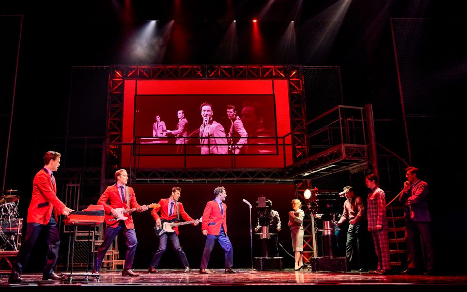 Four men in red jackets playing music with a screen above them