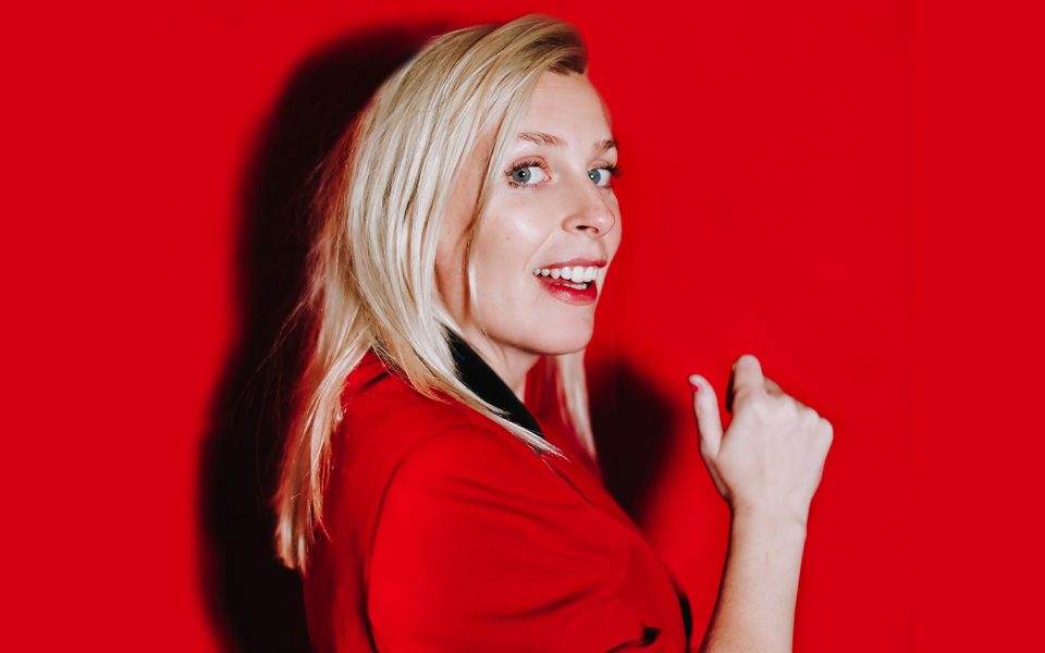 Sara Pascoe wearing a red blazer and red lipstick, smiling against a red background