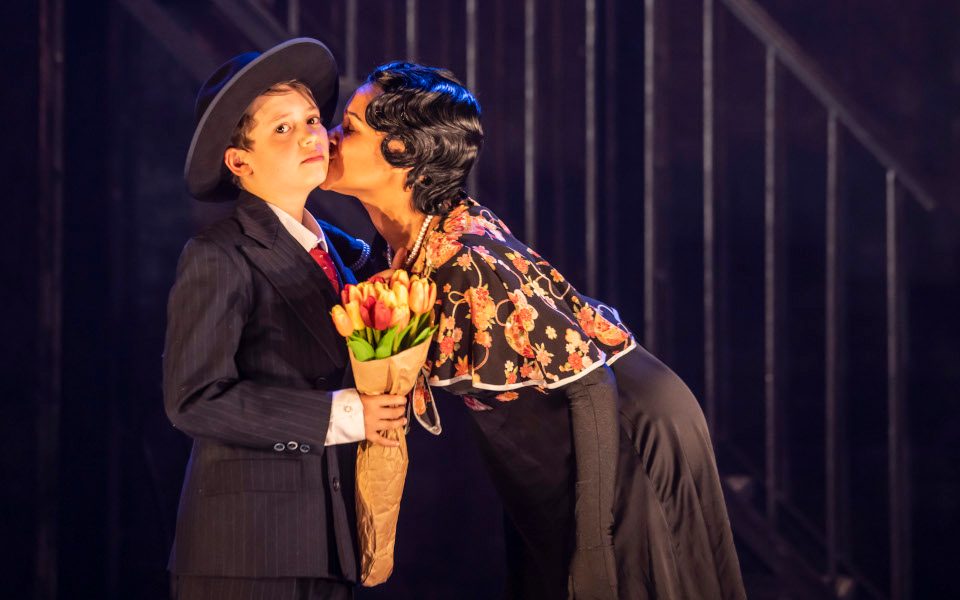 A young person in a suit and hat holding a bunch of flowers being kissed on the cheek by a woman with black hair