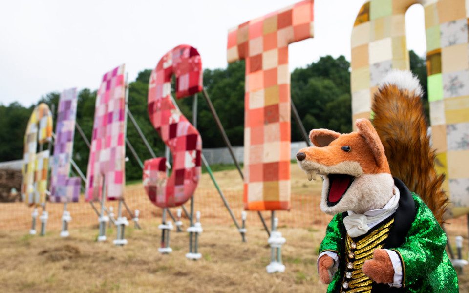 Basil Brush in a green jacket and gold waistcoat in front of giant colourful letters spelling Glasonbury in a field