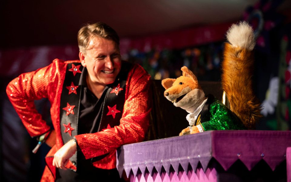 Mr Martin leaning over in a red starry jacket towards Basil Brush on a purple podium