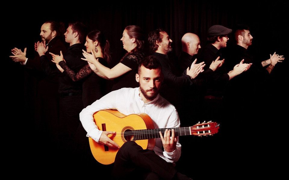 Daniel Martinez at the forefront playing an acoustic guitar, dancers are stood in a line behind