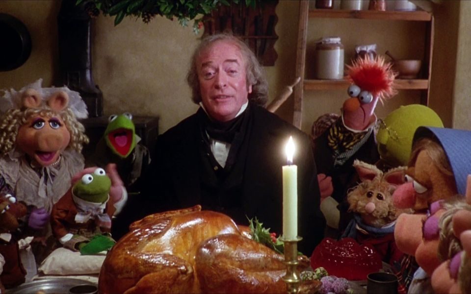 Michael Caine as Ebenezer Scrooge surrounded by various Muppets including but limited to Kermit and Miss Piggy as characters from A Christmas Carol. A big roasted turkey appears on the table in front of them with a candle.