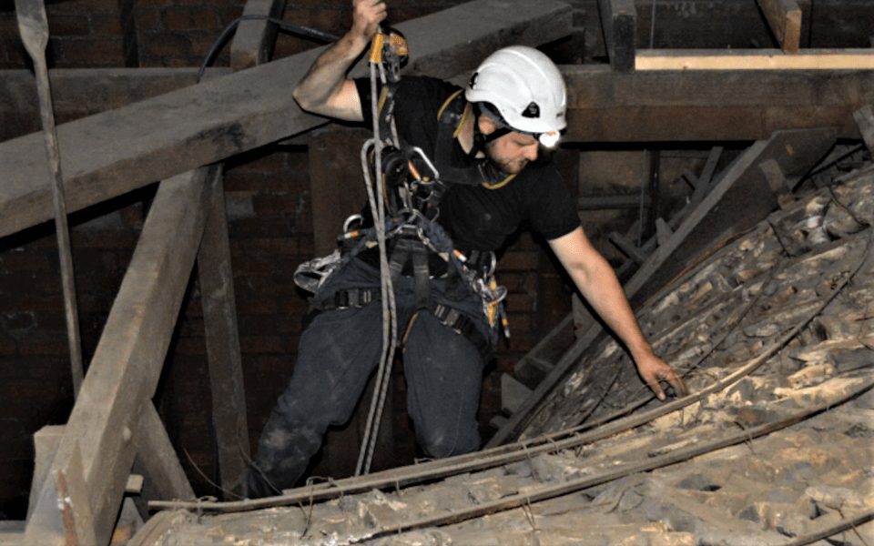 Paul is suspended by a harness and is wearing a hard hat checking the ceiling from above.