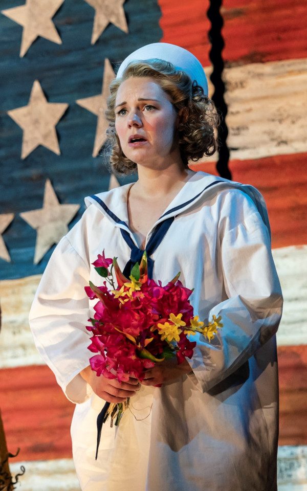 Gina Beck as Nellie Forbush in a white sailor costume holding a bunch of flowers in front of an American flag