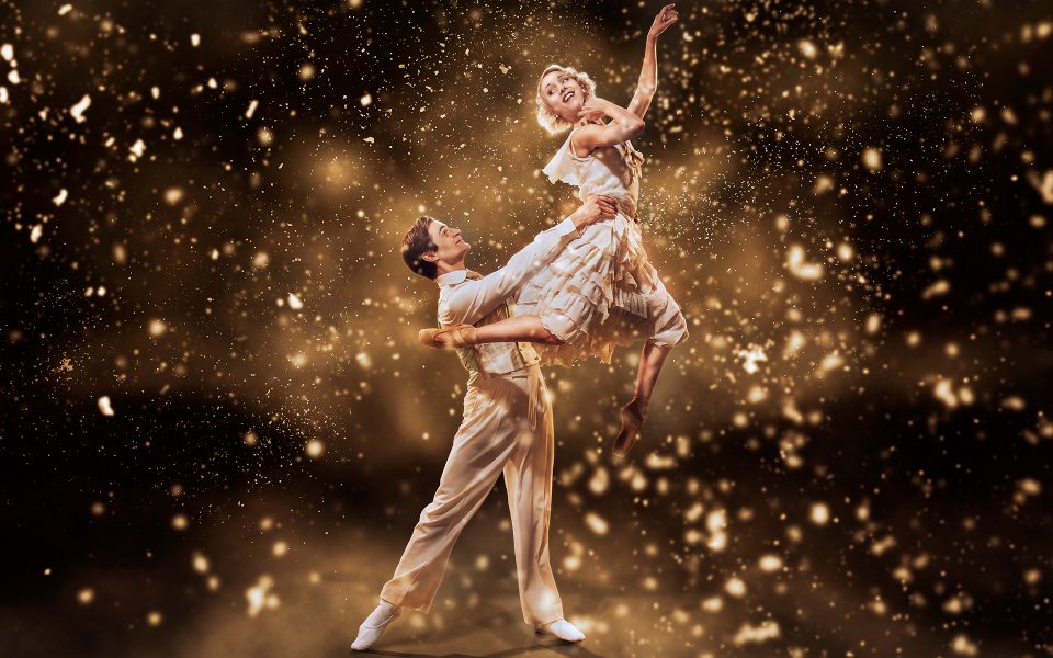 A man in white holding up a woman in white in a ballet pose with a sparkling background