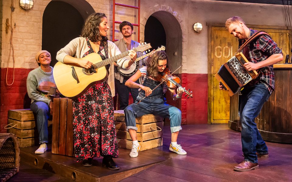 A woman playing the guitar with other musicians around her playing the violin, banjo and accordian