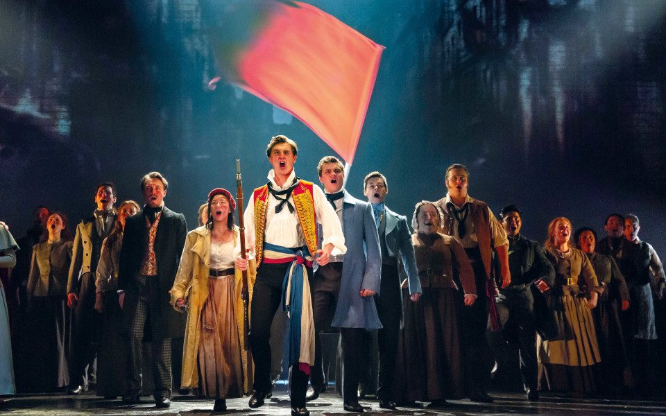 The cast of Les Mis stand in a group singing beneath a red flag waving