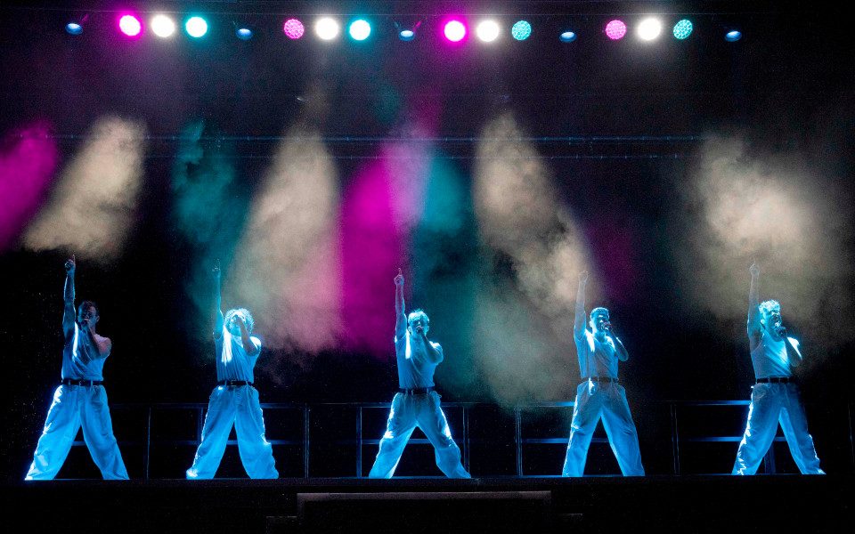 Regan Gascoigne, Kalifa Burton, Archie Durrant, Jamie Corner & Alexanda O'Reilly in Greatest Days. The men, wearing all-white outfits, are singing on stage and dancing in unison.