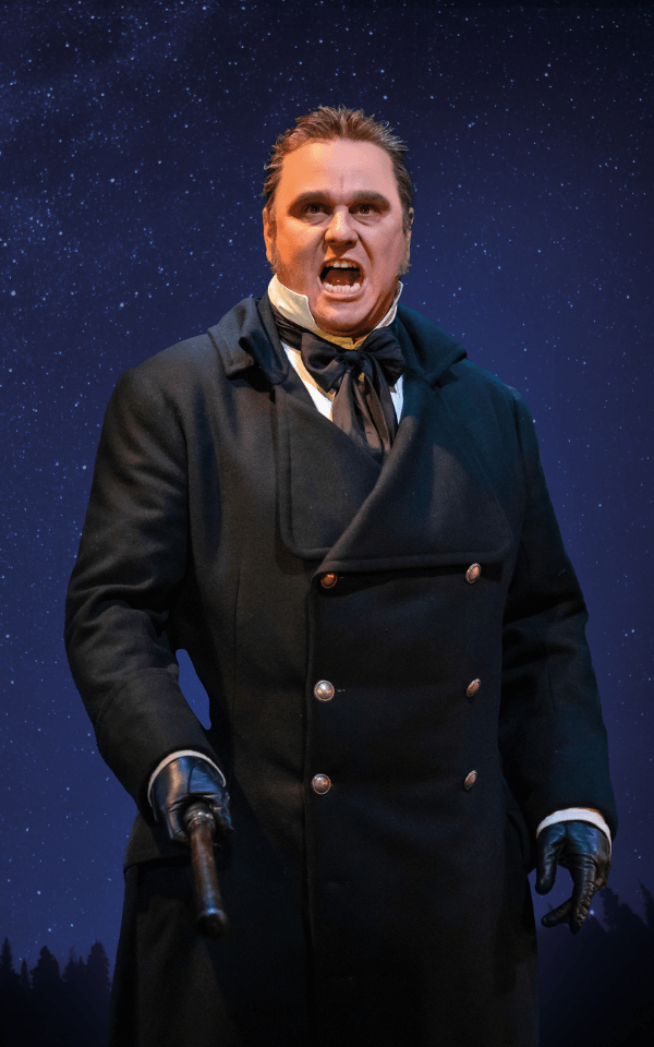 Nic Greenshields as Javert in a black trench coat holding a baton