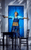 MAMMA MIA! A woman in blue overalls and a utility belt holding a drill with her back to a blue door looking shocked