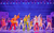 MAMMA MIA! Three woman in colourful 1980s style flared jumpsuits singing into microphones and pointing up. They are surrounded by backing dancers