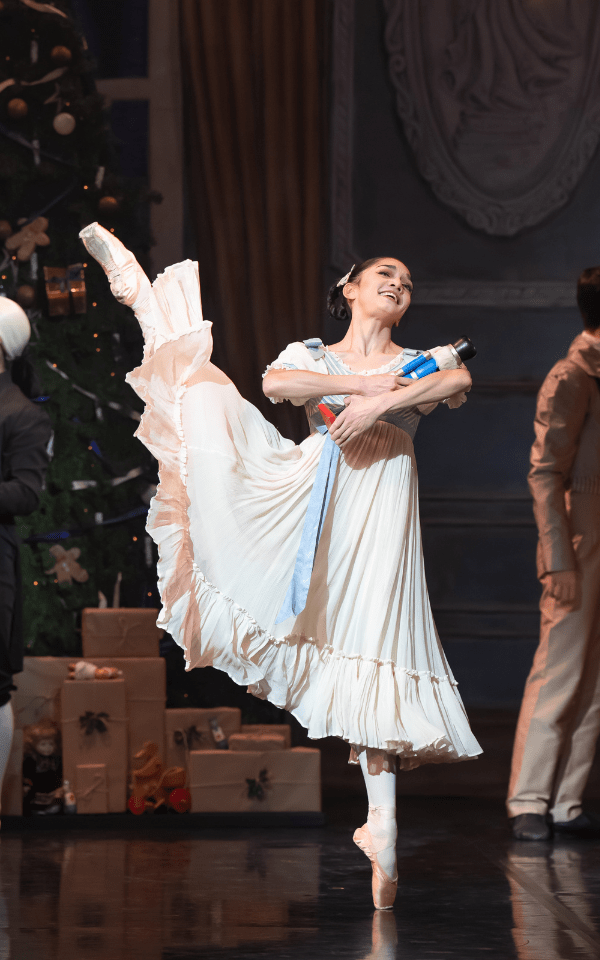 A ballerina in a long light pink dress with her leg in the air, holding a nutcracker