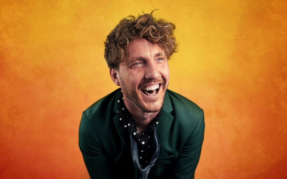 Seann Walsh laughing wearing a green suit in front of orange background