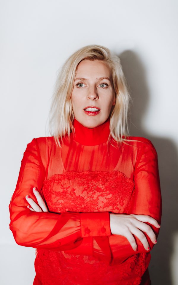 Sara Pascoe smiling wearing a red suit outfit and red lipstick