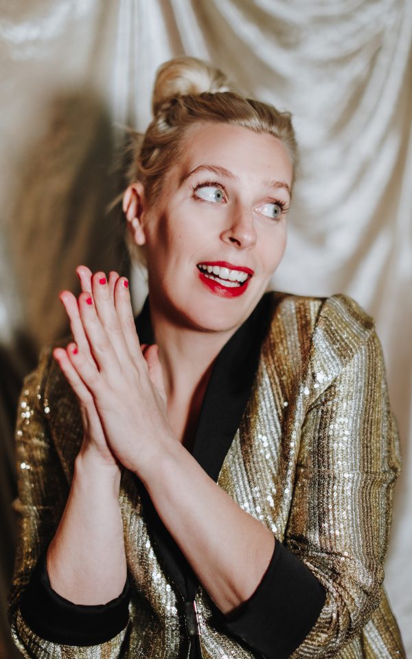 Sara Pascoe smiling wearing a gold suit jacket and red lipstick