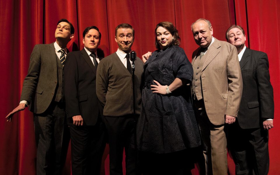 Full Cast of Hancock's Half Hour. Everyone is dressed up in professional attire standing in front of a microphone. There is a red curtain backdrop.