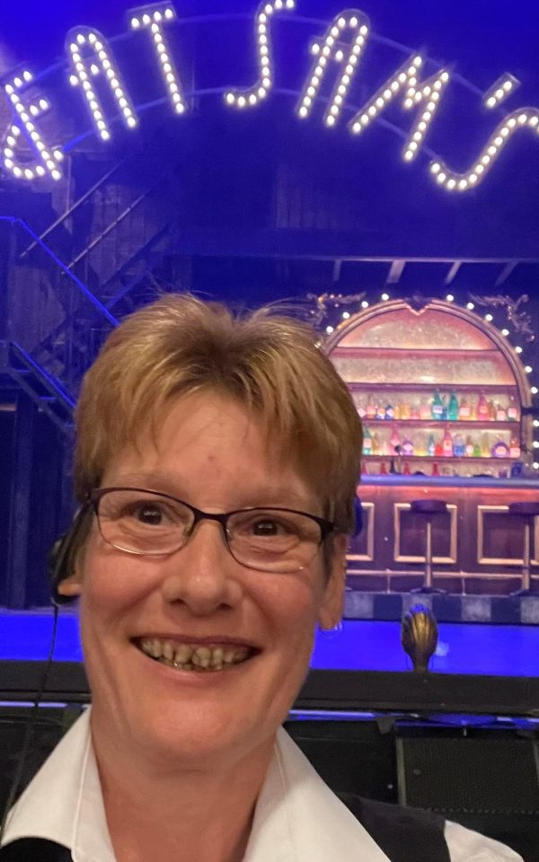 Jaki Taylor in front of the stage at The Grand with the bar from the Bugsy Malone set in the background. Bar is brightly lit with different coloured bottles.
