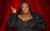 Judi Love Judi Love is in a black outfit with flower petals. She is pointing up and looking to the side with a knowing look. She stands in front of a red curtain background.