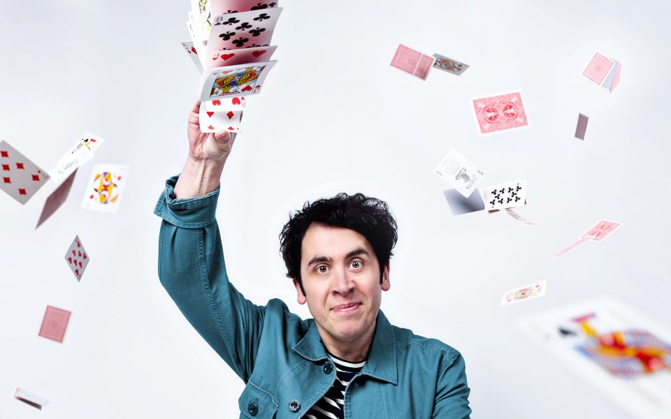 Pete Firman in a striped shirt with a jean jacket overtop. He has playing cards in his hands and is beginning to toss them into the air.