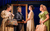 Pride and Prejudice* (*sort of) A woman in a Regency-period suit jacket stands behind a picture frame held up by a woman in a Regency-period dress. Two women holding large bags look at the woman in the picture frame.