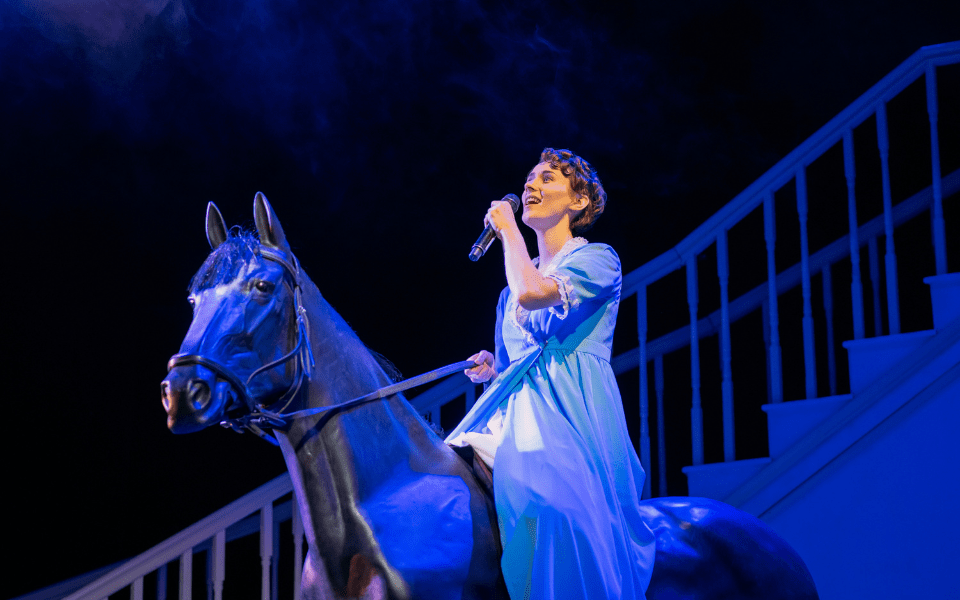 A woman in a blue dress sits on a fake horse and sings into a microphone. They are in blue lighting.