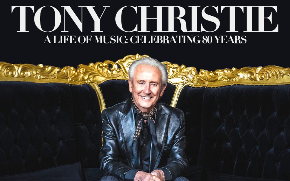 Tony Christie is in a leather jacket with striped trousers. He is smiling while sitting on a velvet couch with gold trim. The background is black.
