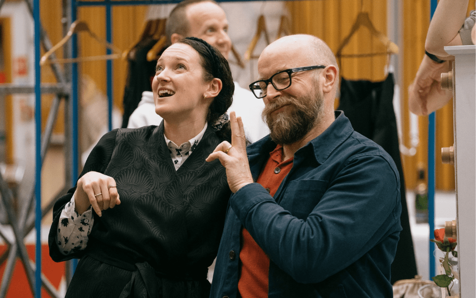 A woman looks up and next to her a man in glasses holds his finger up to her