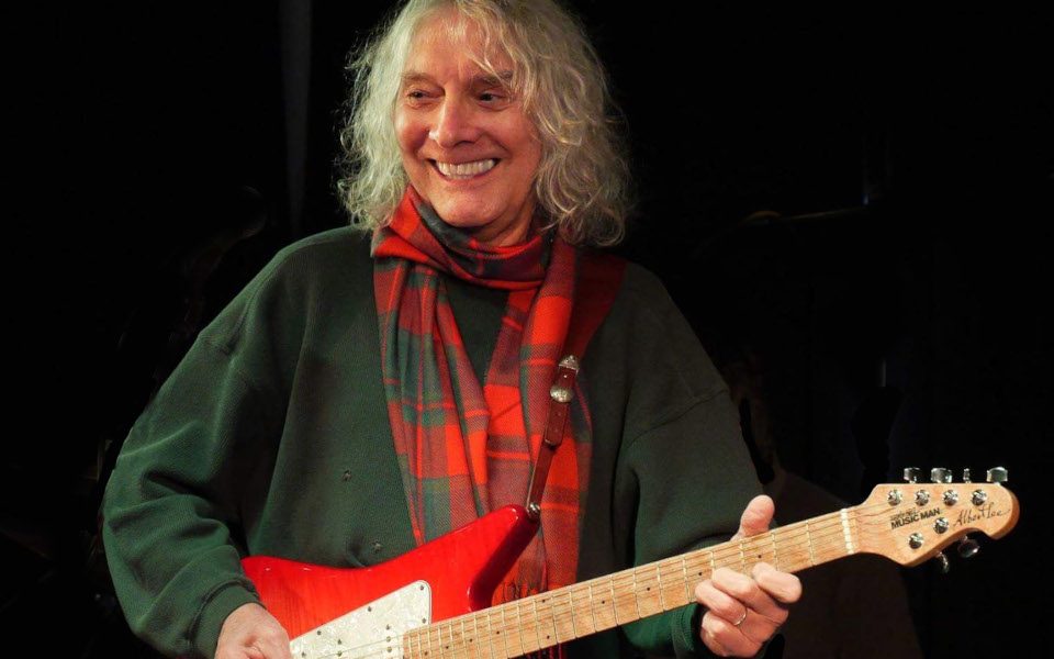 Albert Lee is wearing a red plaid scarf and holding a red electric guitar. He is smiling off to the side.