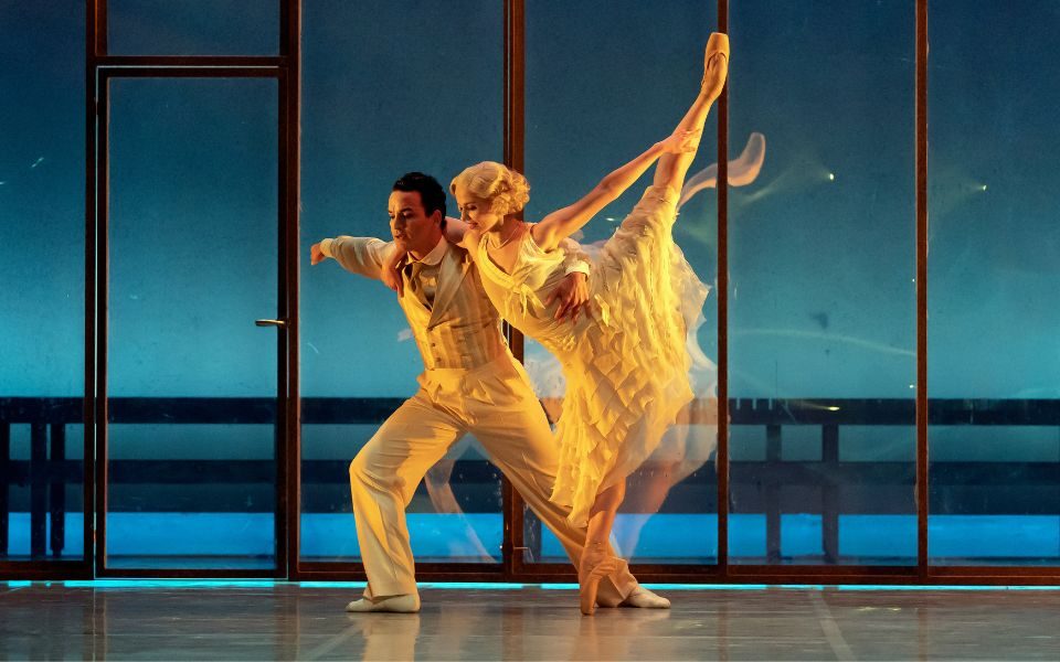 A man and woman have their arms around each others' shoulders and lean forward in a ballet pose. The woman has her leg straight up in the air as she stands on pointe.